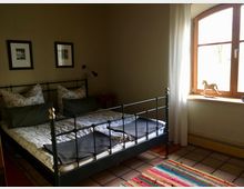 Schlafzimmer mit einem schwarzen Metallbett, floral gemusterter Bettwäsche und zwei Bildern an der Wand. Rechts befindet sich ein großes Fenster mit Holzrahmen, einer weißen Gardine und einem kleinen Schaukelpferd als Dekoration auf der Fensterbank.