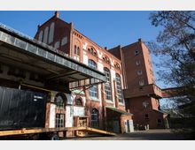 Backsteingebäude eines ehemaligen Industriekomplexes mit hohen Fenstern und verzierten Fassadenelementen. Im Vordergrund ein überdachter Ladebereich und eine schwarze Frachtbox, links und rechts von Bäumen umgeben.