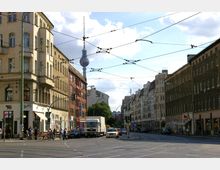 Straßenszene in Berlin mit mehrstöckigen Wohn- und Geschäftshäusern auf beiden Seiten, Straßenbahndrähte über der Kreuzung und dem Fernsehturm im Hintergrund. Autos, Fahrräder und Fußgänger sind im urbanen Umfeld sichtbar.
