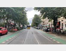 Straßenszene mit einer gelben Straßenbahn in der Mitte, flankiert von Bäumen, geparkten Autos, Fahrrädern und Geschäften auf beiden Seiten. Die Straße hat grüne Fahrradwege und ist von historischen Gebäuden mit Cafés und Läden gesäumt.