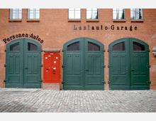 Roter Backsteinbau mit drei großen grünen Toren und einem kleinen roten Eingang in der Mitte, darüber die Beschriftungen „Personen-Autos“ und „Lastauto-Garage“. Der Boden besteht aus Kopfsteinpflaster.