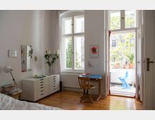 Helles Zimmer mit Holzboden, einem weißen Schrank, einem kleinen Schreibtisch mit Holzstuhl und einer Balkontür, die zu einem Balkon mit Pflanzen und einem blauen Stuhl führt. Große Fenster lassen Tageslicht herein, und an der Wand hängen minimalistische Dekorationen.