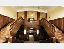 Holzvertäfelter Eingangsbereich eines historischen Gebäudes mit einer symmetrischen Doppeltreppe, die zu einem oberen Zwischenpodest führt. Große, verglaste Holztüren und Wandlampen rahmen den unteren Bereich ein.