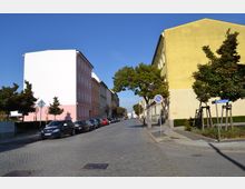 Straßenszene in einer Wohngegend mit gepflasterter Fahrbahn, links ein rosa und rechts ein gelbes Gebäude. Entlang der Straße sind geparkte Autos und junge Bäume mit Stützen zu sehen; ein Straßenschild zeigt die "Albrechtstraße".