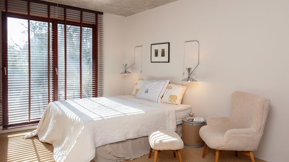 Minimalist bedroom with a double bed dressed in white linens, a raw concrete ceiling, and a large sliding glass door with horizontal blinds letting in strong sunlight. A small side table, wall-mounted reading lights, and a cushioned chair with matching ottoman sit against a plain white wall on a neutral carpet.
