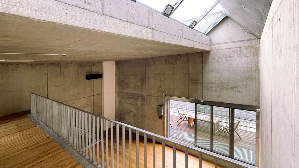 Interior of a modern concrete building with a wooden floor walkway and metal railing overlooking a lower level. Large skylights and a wall of sliding glass doors open to a small outdoor deck with seating.