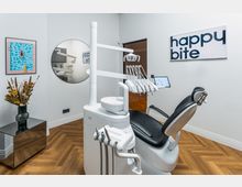 Moderner Behandlungsraum in einer Zahnarztpraxis mit einem weißen Zahnarztstuhl und zahnmedizinischen Geräten. Der Raum hat einen Holzboden, ein rundes Wandspiegel, dekorative Trockenblumen auf einem Tisch und eine helle Wand mit Kunstbildern und dem Schriftzug "happy bite".