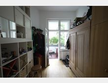 Schmaler Flur mit Holzfußboden, auf der linken Seite ein weißes Regal mit offenen und geschlossenen Fächern, daneben eine Garderobe mit Jacken. Auf der rechten Seite ein großer Holzschrank sowie Schuhe auf einer Ablage. Am Ende des Raumes eine Glastür, die zu einem kleinen Balkon mit Gartenblick führt.