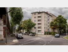 Straßenecke in einem urbanen Wohngebiet mit einem mehrstöckigen Wohnhaus aus Beton, mehreren Bäumen und parkenden Autos am Straßenrand. Im Vordergrund ist ein Straßenschild mit der Aufschrift „Alt-Moabit“ zu sehen.