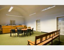 Ein schlichter Gerichtssaal mit grüner Teppichboden, einer hölzernen Richterbank im Hintergrund, mehreren Tischen und schwarzen Stühlen in der Mitte sowie einer Holzbank im Vordergrund. An den Wänden befinden sich zwei cremefarbene Türen mit quadratischem Muster.