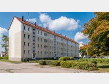 Mehrstöckiges Wohngebäude mit heller Fassade, rotem Satteldach und zahlreichen Fenstern, umgeben von gepflegten Hecken und einem Baum rechts im Vordergrund; im Hintergrund blauer Himmel mit Wolken.