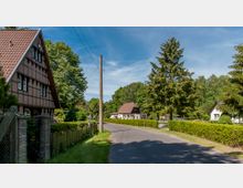 Eine ländliche Straße mit einem traditionellen Fachwerkhaus links und mehreren Einfamilienhäusern mit Satteldächern im Hintergrund, umgeben von hohen Bäumen und gepflegten Hecken. Der Himmel ist klar und blau, und ein Strommast steht am Straßenrand.