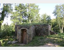 Ein kleines, rechteckiges Gebäude aus Naturstein und Ziegelmauerwerk mit einer Holztür in der Mitte, umgeben von Bäumen und Grünflächen in einer ländlichen, bewaldeten Umgebung. Das Dach ist mit Gras bewachsen, was auf eine frühere Nutzung als Eiskeller oder Lagerraum hindeuten könnte.