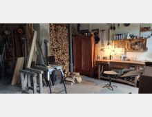 Eine Werkstatt mit Holzstapeln an einer Wand, einer Holzwerkbank mit Werkzeugen und Beleuchtung, sowie einem Ofenrohr. Im Raum befinden sich verschiedene Holzplatten, Geräte und ein alter Schrank, neben einem Tisch und einem Drehstuhl.