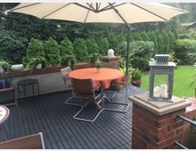 Garten-Terrasse mit Holzboden, einem runden Tisch mit orangefarbener Tischdecke und Stühlen unter einem großen Sonnenschirm. Im Hintergrund sind immergrüne Sträucher und eine Rasenfläche zu sehen, auf der Terrasse stehen dekorative Elemente wie eine Metalllaterne und Blumentöpfe.