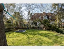 Grüne Gartenlandschaft mit einer Rasenfläche, einem eingelassenen Trampolin und einer Slackline, die zwischen Bäumen gespannt ist. Im Hintergrund ein weißes Haus mit roten Dachziegeln und Dachgauben, umgeben von Bäumen und Sträuchern.