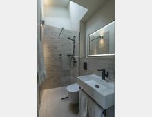 Modernes Badezimmer mit schlichter Einrichtung, einer Glasdusche mit schwarzem Duschkopf und beiger Fliesenwand. Neben dem Waschbecken mit schwarzem Wasserhahn und beleuchtetem Spiegel befindet sich eine schwebende Toilette, und Tageslicht fällt durch ein Oberlicht ein.