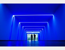 Ein moderner, blau beleuchteter Gang mit geometrischen Lichtstreifen an Decke und Wänden. Im Hintergrund sind drei Personen zu sehen, zwei in der Mitte in Silhouette und eine entfernte Person am Ende des Gangs vor einem helleren Bereich.