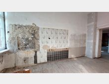 Ein leerer Raum mit stark beschädigten Wänden, teilweise mit freigelegtem Mauerwerk, alten weißen Fliesen mit floralem Muster und schwarzen Kacheln unten. Ein Fenster links lässt Tageslicht herein, und rechts ist eine geöffnete Tür sichtbar.