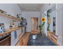 Helle Küche in einem Altbau mit Stuckdecke, langen Holzarbeitsflächen, offenen Regalen und einer modernen Einrichtung. Im Vordergrund steht ein Tisch mit einer Sonnenblume in einer Vase, im Hintergrund ist eine offene Tür zu einem angrenzenden Raum zu sehen.