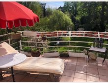 Terrasse mit terrakottafarbenen Fliesen, einem Liegestuhl, einem kleinen Marmortisch und einem roten Sonnenschirm. Im Hintergrund ist ein Garten mit Rasen, blühenden Büschen und hohen Bäumen zu sehen, während die Balkonbrüstung mit Blumentöpfen dekoriert ist.