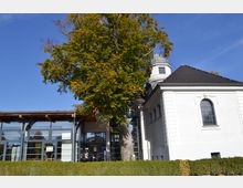 Ein Gebäudeensemble mit einem weißen, historisch anmutenden Bau mit Rundbogenfenster und dunklem Schieferdach, daneben ein moderner Anbau mit großen Glasfronten und Holzelementen. Ein großer Baum mit herbstlichem Laub steht zentral vor den Gebäuden, umgeben von einer niedrigen Buchsbaumhecke.