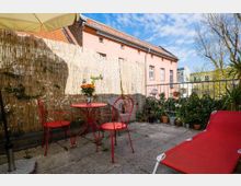 Eine kleine Terrasse mit Steinfliesen, einem roten Metalltisch und passenden roten Stühlen. Im Hintergrund eine Bambusmatte als Sichtschutz, mehrere Topfpflanzen entlang eines Geländers und Fassaden von Wohngebäuden, darunter ein pinkfarbenes Haus.