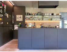 Moderne Küche mit einer Front aus matten, grauen Schränken, einer Betonwand mit offenen Regalböden, auf denen Geschirr und Gläser stehen, und einer Arbeitsplatte mit Küchengeräten und einem Kochfeld. Rechts hängt eine schwarze Dunstabzugshaube, links ist eine Wand mit dunklen Magneten und Dekorationen.