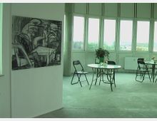 Ein heller Raum mit großen Fenstern, die eine Aussicht auf eine Stadtlandschaft bieten. Im Vordergrund steht ein Tisch mit Stühlen, dekoriert mit einer Vase und einer Pflanze; an der Wand hängt ein großformatiges Schwarz-Weiß-Gemälde.