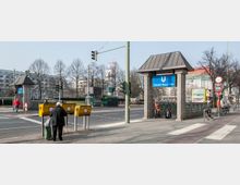 Stadtszene mit dem Eingangsbereich der U-Bahnstation „Theodor-Heuss-Platz“ in Berlin, erkennbar an einem blauen Schild mit weißer Schrift. Im Vordergrund befinden sich gelbe Briefkästen der Deutschen Post, ein Gehweg und eine Straße mit Ampeln; im Hintergrund stehen Bäume und städtische Gebäude.