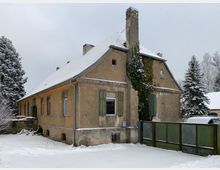 Schneebedecktes, älteres Gebäude mit Putzfassade, grünen Fensterläden und Schornstein, teils von Efeu bewachsen. Es steht in einer winterlichen Landschaft mit schneebedecktem Boden und umliegenden Bäumen.