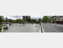 Panorama eines urbanen Platzes mit breiten Gehwegen, links flankiert von Bäumen und Cafés mit roten Sonnenschirmen, im Hintergrund die Ruine der Kaiser-Wilhelm-Gedächtniskirche. Rechts verläuft eine Straße mit Pollern und angrenzenden modernen Gebäuden mit Geschäften im Erdgeschoss.