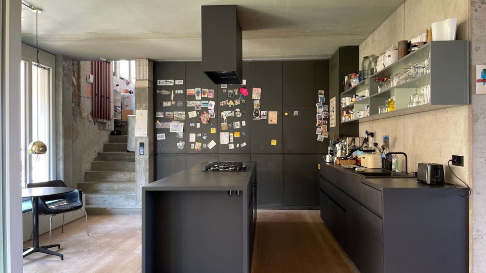 Moderne Küche mit dunklen Einbauschränken, einer zentralen Kochinsel und offenen Regalen an einer Betonwand. Im Hintergrund führt eine Betontreppe zu einem oberen Bereich, und links befindet sich eine Glasfront mit Blick nach draußen.