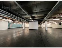 Großer, leerer Innenraum eines Keller- oder Industriegebäudes mit Betonboden, sichtbarer Betondecke und freigelegten Lüftungsrohren sowie Beleuchtungskörpern. Weiße Wände und vereinzelte Säulen unterteilen den Raum, der spärlich beleuchtet ist.