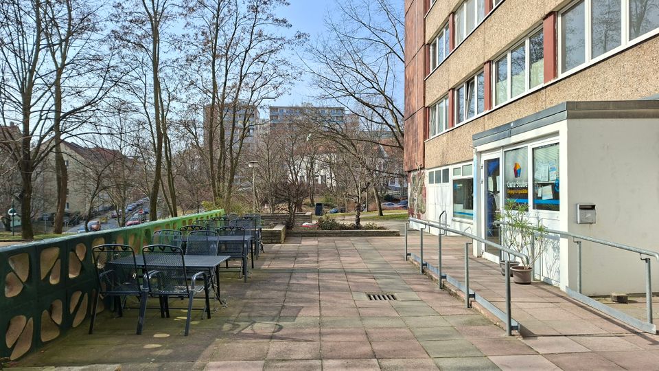 Terrasse vor einem mehrstöckigen Gebäude mit großen Fenstern, einem Eingang mit Glastür und einer Rampe mit Geländer. Links stehen mehrere Metalltische und -stühle entlang einer grünen Brüstung; im Hintergrund sind Bäume, Wohnhäuser und eine Straße zu sehen.