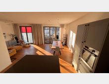 Wohnzimmer mit offener Küche und Holzboden, gestaltet mit modernen Möbeln. Helle Fensterfront mit Vorhängen, ein Esstisch mit Stühlen steht im Mittelpunkt, und links ist eine Sitzecke mit Sofa und Sessel sichtbar.