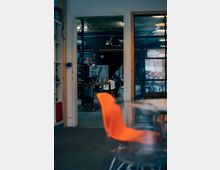 Ein Werkstatt- oder Bürobereich mit offenen Türen zeigt einen Raum im Hintergrund, in dem Werkzeuge und Geräte sichtbar sind. Im Vordergrund stehen orangefarbene und transparente Stühle vor einem Tisch.
