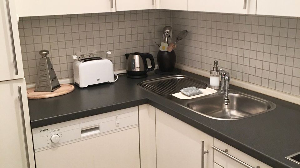 Eckküche mit weißen Schränken und dunkler Arbeitsplatte, grauer Fliesenrückwand und Edelstahlspüle mit Abtropffläche. Auf der Arbeitsfläche stehen Küchenutensilien wie Toaster, Wasserkocher und Reibe; unten sind Geschirrspüler und Schubladen integriert.