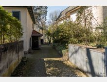 Schmaler gepflasterter Weg in einem Wohnbereich, flankiert von einem weißen Haus mit grünen Fensterläden auf der linken Seite und einer Hecke sowie Metallbehältern auf der rechten Seite. Im Hintergrund sind Bäume und ein weiteres Gebäude zu sehen.