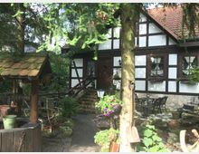 Fachwerkhaus mit weißen Wänden und dunkelbraunen Holzbalken, umgeben von einem Garten mit Bäumen, Blumen und rustikalen Dekorationen wie einem Brunnen mit Holzdach und einem alten Spaten. Der Eingang ist über eine kleine Steintreppe erreichbar, und eine Terrasse mit Metallmöbeln ist sichtbar.