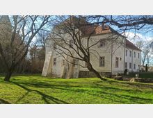 Historisches Gebäude mit heller Putzfassade, roten Ziegeldach und mehreren kleinen Fenstern, umgeben von einem grünen Rasen mit vereinzelten kahlen Bäumen. Im Vordergrund stehen Treppenstufen und dekorative Steinvasen, während im Hintergrund Sträucher und ein klarer Himmel zu sehen sind.