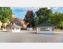 Weitläufiger Platz mit mehreren weißen Pavillons mit dunklen Dächern, umgeben von Bäumen und einem backsteinernen Gebäude mit großen Fenstern im Hintergrund. Der Himmel ist teils bewölkt, und die Umgebung wirkt offen und ruhig.