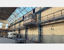 Große Industriehalle mit hohen Backsteinwänden, Stahlträgern und einem durchgehenden Glasdach, das viel Tageslicht hereinlässt. Entlang der Wand verlaufen Rohrleitungen und eine erhöhte Laufbahn; am Boden stehen ein Wohnmobil, ein Tischtennistisch und einige Stühle.