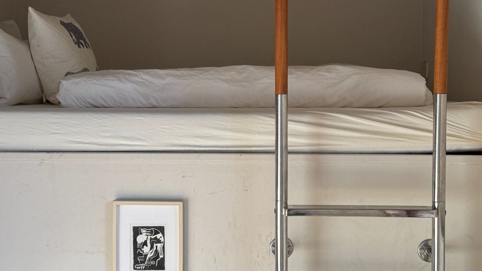Loft-style sleeping area with a low concrete ceiling, featuring a bed with white bedding and a wall-mounted metal ladder with wooden handrails leading up to it. A plain white wall below includes a small framed artwork.