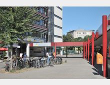 Außenbereich eines Bürogebäudes mit einem Schild der Deutschen Rentenversicherung, Fahrradständern im Vordergrund und roten Pfeilern eines Vordachs rechts. Im Hintergrund sind weitere Gebäude und ein kleiner Ladenbereich erkennbar.