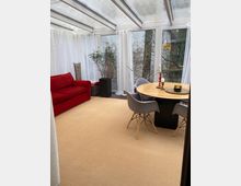 Wohnbereich mit gläsernem Wintergartendach und großen Fenstern, die von weißen Vorhängen eingerahmt sind. Der Raum ist mit einem roten Sofa, einem runden Holztisch mit grauen Stühlen und mehreren Pflanzen dekoriert.