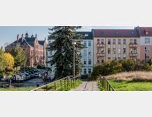 Ein Fußweg mit Geländern führt durch eine grüne Wiese, im Hintergrund stehen moderne Wohnhäuser und ein großes rotes Backsteingebäude mit Türmchen. Dazwischen sind Bäume, parkende Autos und eine Straßenkreuzung zu sehen.