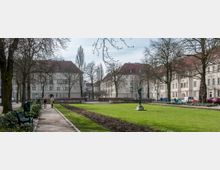 Ein ruhiger Platz mit einer Grünfläche, umgeben von mehrstöckigen Wohngebäuden mit roten Dächern und heller Fassade. Im Vordergrund führen Wege entlang von Bäumen und Bänken, während eine bronzene Statue auf der Wiese steht.