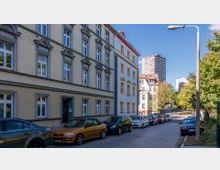 Straßenansicht in einer Stadt mit historischen Wohngebäuden, die helle Fassaden und dekorative Fensterrahmen haben. Autos parken entlang der schmalen gepflasterten Straße, und im Hintergrund sind moderne Hochhäuser sowie Bäume erkennbar.