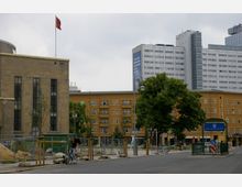 Straßenszene am Rosa-Luxemburg-Platz in Berlin mit einem historischen Gebäudeteil links, modernen Hochhäusern im Hintergrund und einem gelb-orangefarbenen Wohnhaus in der Mitte. Auf der rechten Seite ist der U-Bahn-Eingang "Rosa-Luxemburg-Platz" zu sehen, umliegende Baustellen sind mit Zäunen abgesichert.